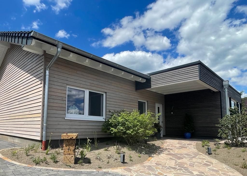 Modernes Einfamilienhaus in Holzbauweise im Kreis Ahrweiler mit heller Holzfassade, überdachtem Eingang und gepflegtem, minimalistischem Vorgarten unter blauem Himmel.