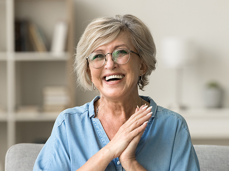 Fröhliche ältere Frau mit perfekten weißen Zähnen dank Keramikkronen sitzt entspannt auf Sofa und lächelt.