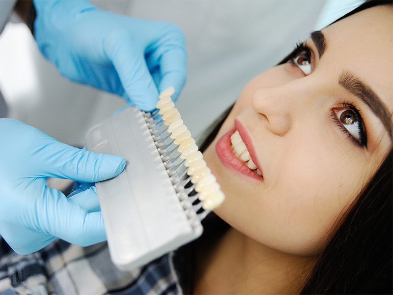 Zahnärztin hält Farbschlüssel an Zähne einer Patientin, um passenden Zahnersatz farblich abzustimmen.